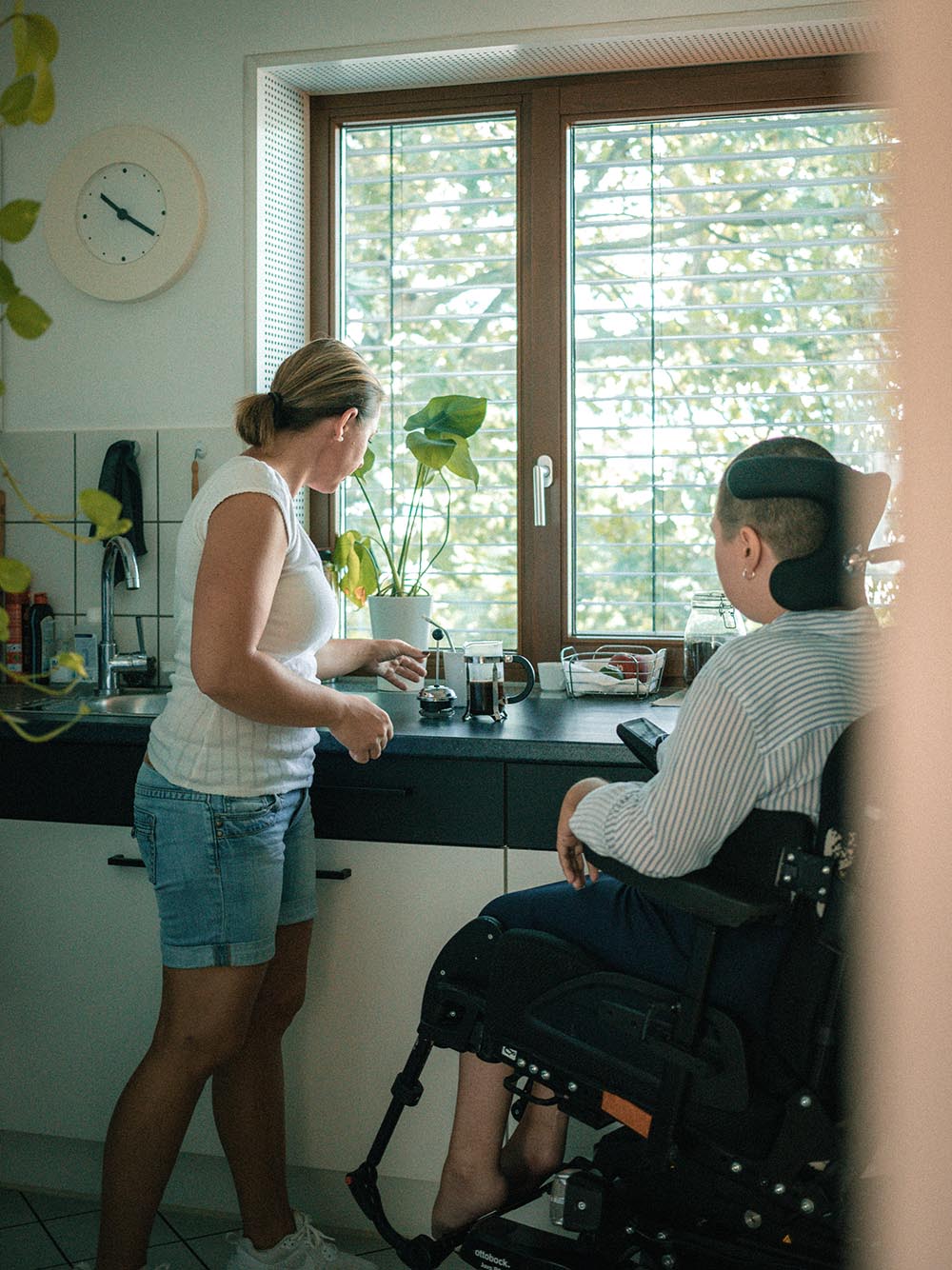 Person sitzt im Rollstuhl steht neben einer anderen Person in der Küche, die stehende Person assistiert, indem sie Kaffee zubereitet.